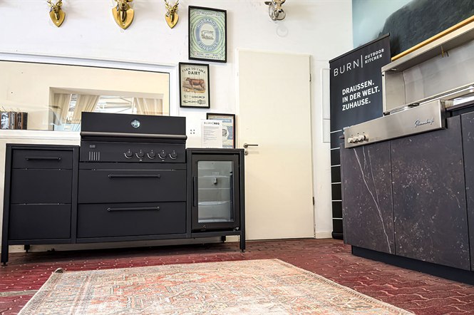 Schwarze Outdoorküche mit Einbaugrill und Kühlschublade, daneben Burn Outdoorküche mit Arbeitsplatte in Marmoroptik
