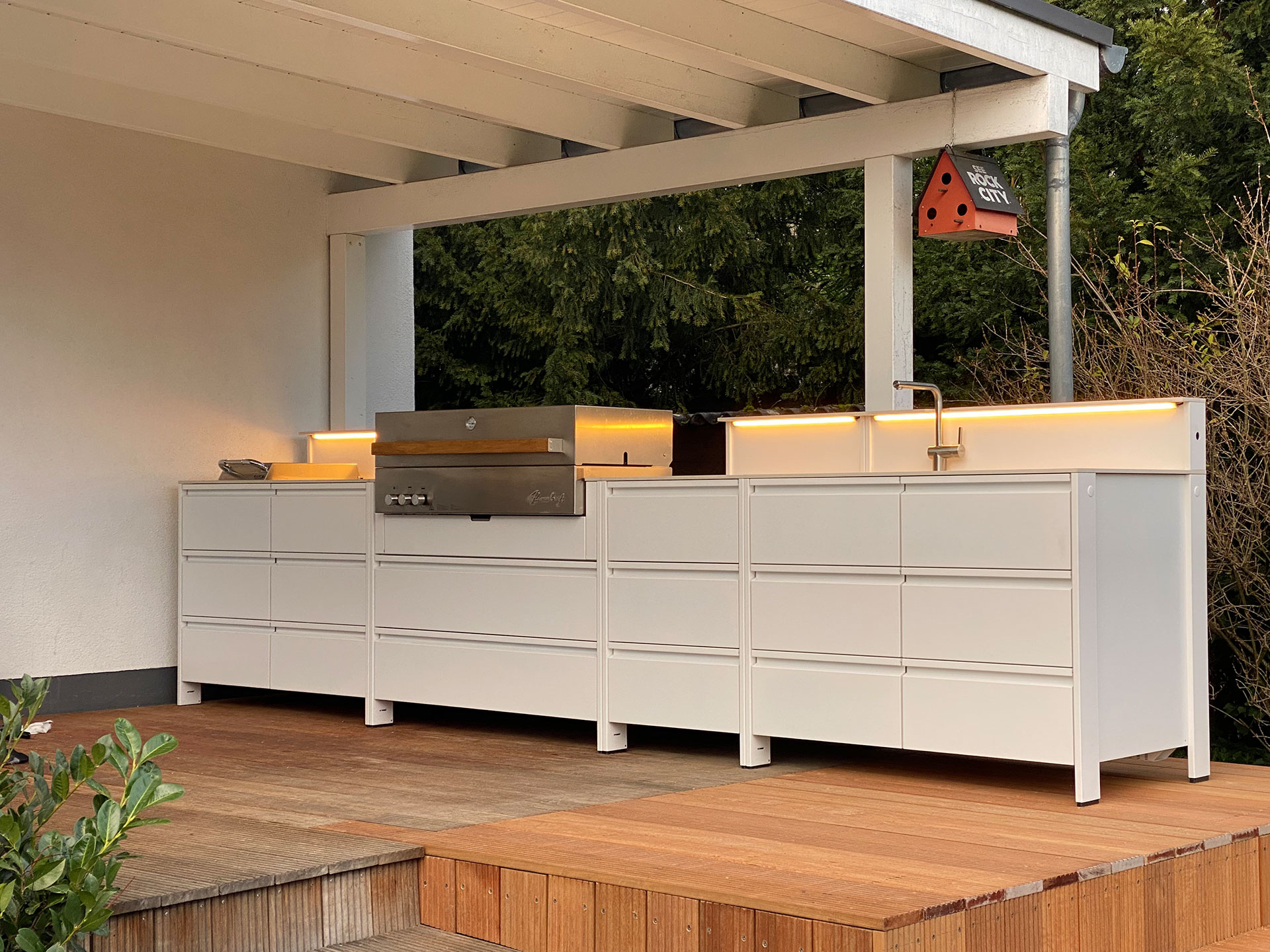 Weiße Belmento Outdoorküche mit integriertem Grill, Spüle und LED-Beleuchtung auf überdachter Holzterrasse im Garten