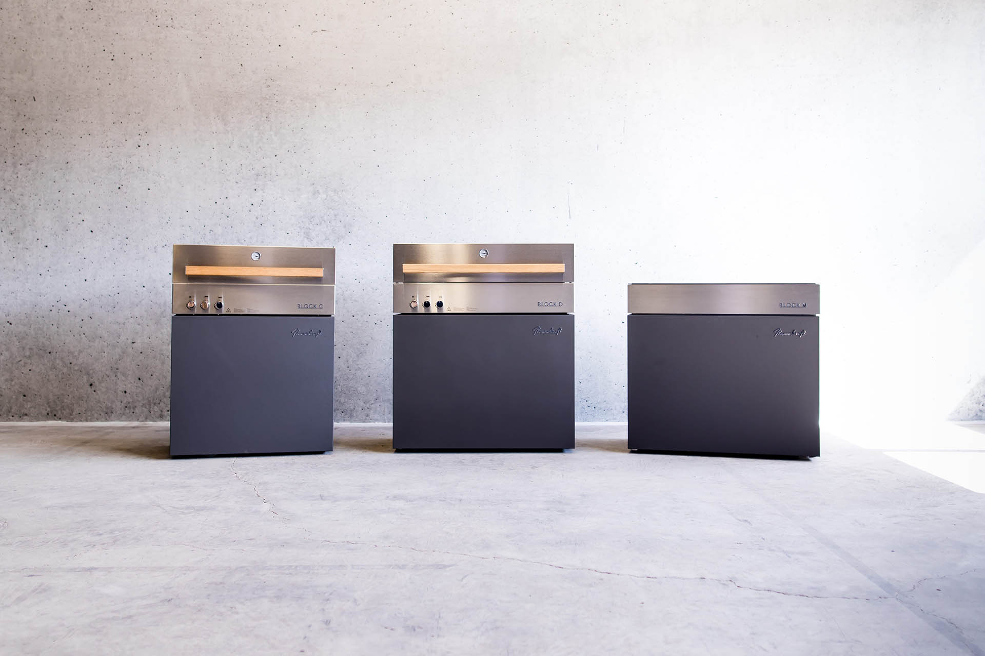 Drei modulare Flammkraft Outdoorküchen-Elemente in modernem Design mit Edelstahlfronten vor minimalistischer Betonwand.