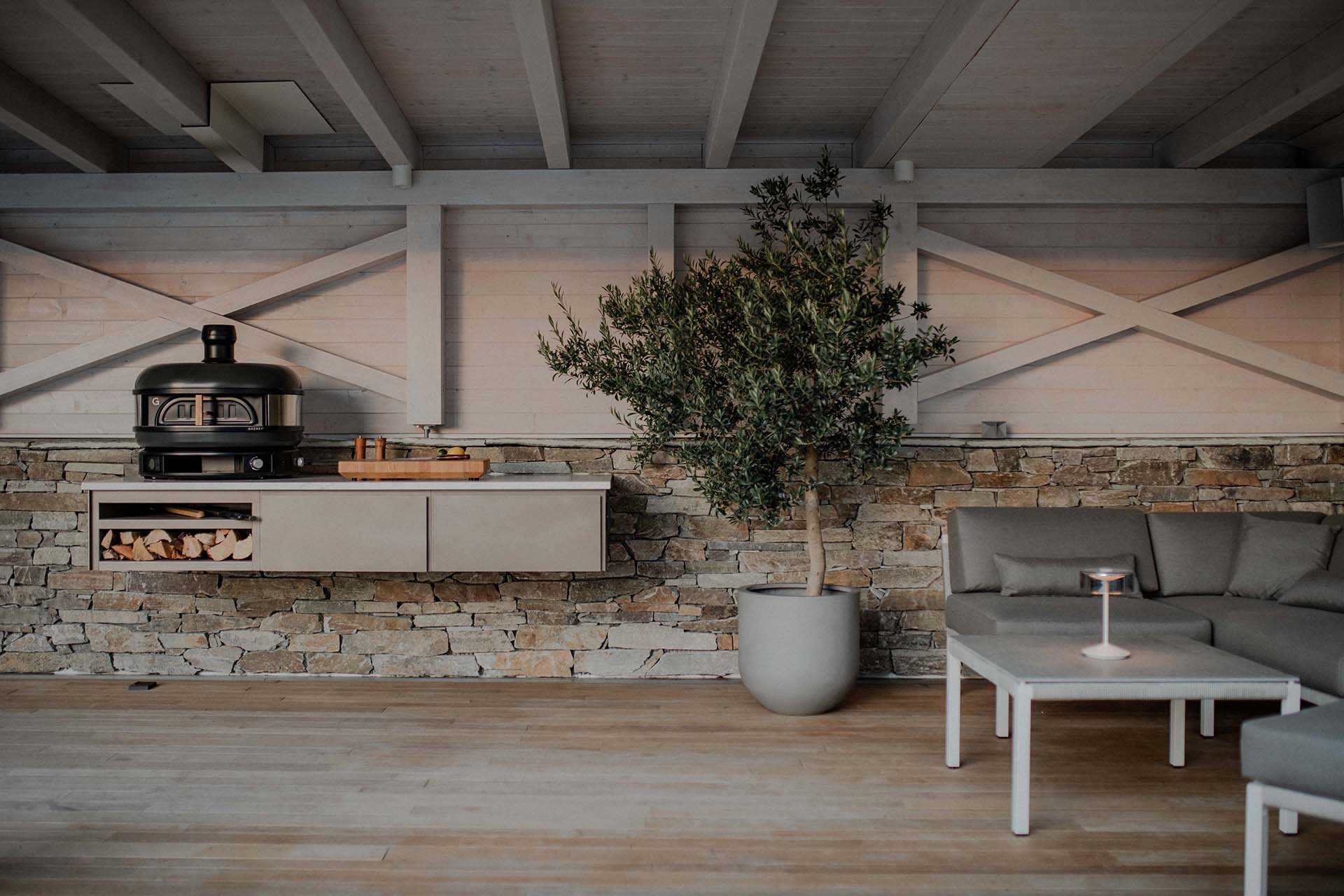 Grillzimmer Outdoorküche mit Pizzaofen und schwebender Modulzeile in stilvoller Lounge-Umgebung mit Natursteinwand und Holzinterieur.